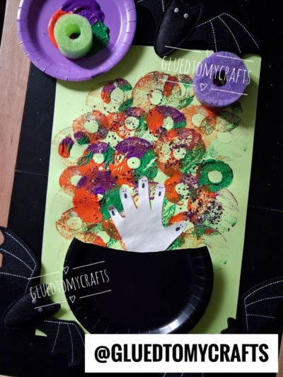 Childrens Halloween craft: Colorful circular sponge prints make bubbles from a black paper cauldron with a white handprint on green paper. Bat decorations and sponges are nearby. Text reads @GLUEDTOMYCRAFTS..