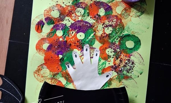 Childrens Halloween craft: Colorful circular sponge prints make bubbles from a black paper cauldron with a white handprint on green paper. Bat decorations and sponges are nearby. Text reads @GLUEDTOMYCRAFTS..