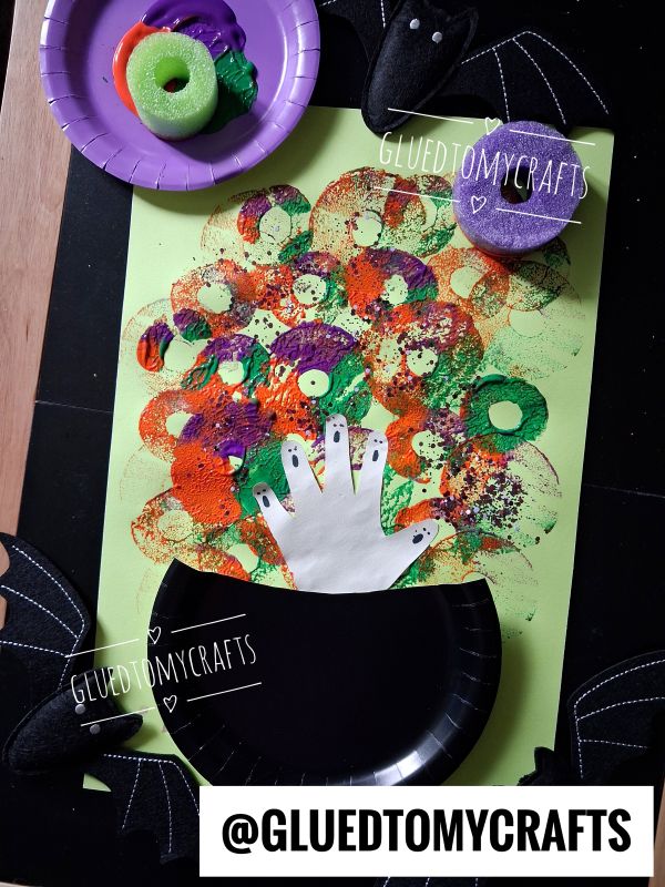 Childrens Halloween craft: Colorful circular sponge prints make bubbles from a black paper cauldron with a white handprint on green paper. Bat decorations and sponges are nearby. Text reads @GLUEDTOMYCRAFTS..