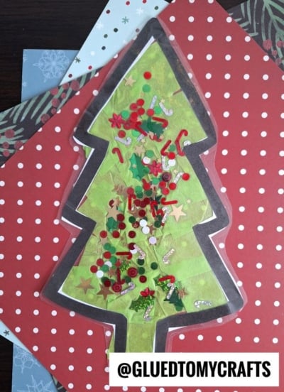 A Christmas tree-shaped craft inspired by a stained glass Christmas tree, adorned with stars, sequins, holly leaves, and candy cane decorations on green paper, set on polka dot and festive patterned backgrounds. GLUEDTOMYCRAFTS is written at the bottom.