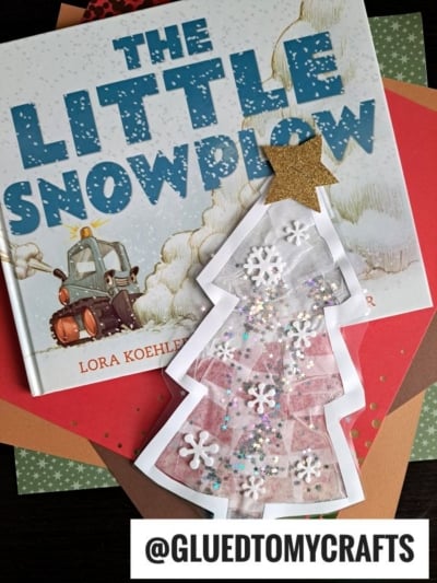 A children’s book titled The Little Snowplow is beneath a stained glass Christmas tree craft decorated with snowflakes, glitter, and a gold star. Colorful paper sheets are layered underneath. Text reads @GLUEDTOMYCRAFTS.