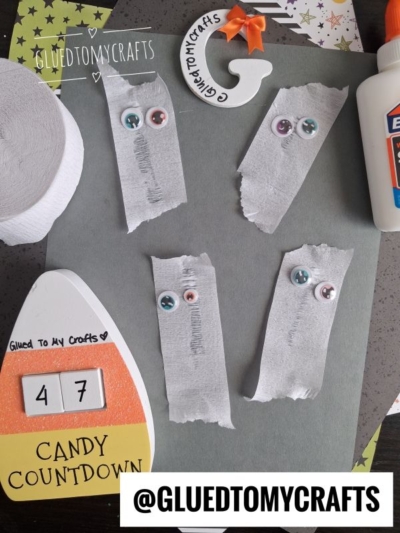 Four tissue paper ghosts with googly eyes are glued to gray paper. Halloween Crafts for Kids, a candy countdown, and a bottle of glue are nearby on a decorated table. @GLUEDTOMYCRAFTS appears twice.