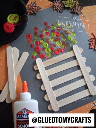 A fall craft project with popsicle sticks arranged on black paper, colorful painted dots, glue, and artificial leaves, with @GLUEDTOMYCRAFTS tag at the bottom.