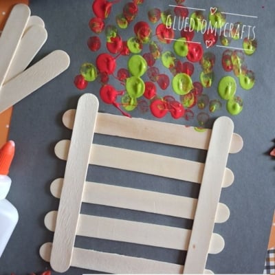 Popsicle stick fence craft on black paper with red, yellow, and green paint dots above, resembling fall leaves. Glue bottle and extra sticks nearby. @gluedtomycrafts watermark on image.