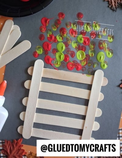 Popsicle stick fence craft on black paper with red, yellow, and green paint dots above, resembling fall leaves. Glue bottle and extra sticks nearby. @gluedtomycrafts watermark on image.
