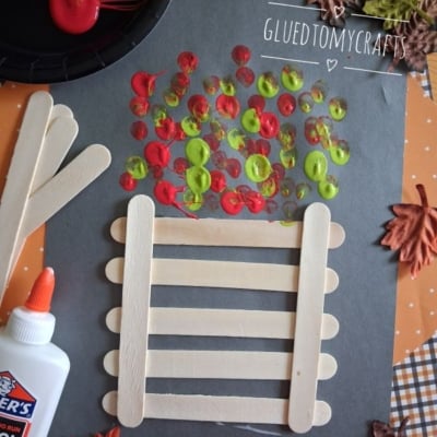 A fall craft project with popsicle sticks forming a fence, red, yellow, and green paint dots as leaves, glue, paint, and artificial autumn leaves on a black paper background.