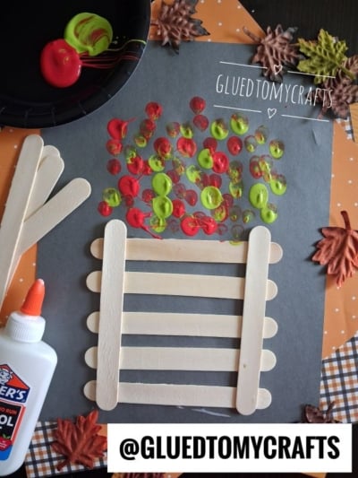 A fall craft project with popsicle sticks forming a fence, red, yellow, and green paint dots as leaves, glue, paint, and artificial autumn leaves on a black paper background.