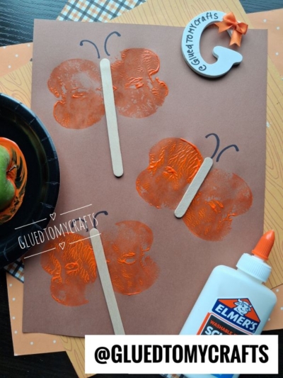 Brown paper with three orange butterfly prints made from an apple stamped craft, each featuring a craft stick as the body. There’s a bottle of glue, a black plate with an apple, and a G pumpkin decoration nearby. @GLUEDTOMYCRAFTS is watermarked.
