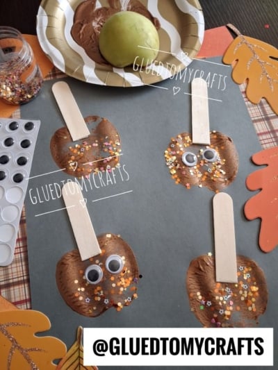 Four apple-themed crafts made from brown paint, craft sticks, googly eyes, and glittery confetti are displayed on gray paper, surrounded by craft supplies and autumn-themed decorations.
