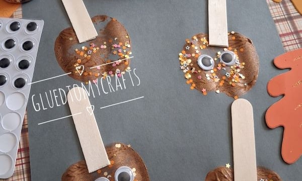 Four apple-themed crafts made from brown paint, craft sticks, googly eyes, and glittery confetti are displayed on gray paper, surrounded by craft supplies and autumn-themed decorations.