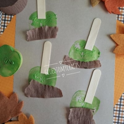 Craft project on gray paper featuring four apple stamped shapes made of green paint, wooden popsicle sticks, and brown crepe paper, surrounded by felt leaves.