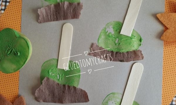 Craft project on gray paper featuring four apple stamped shapes made of green paint, wooden popsicle sticks, and brown crepe paper, surrounded by felt leaves.