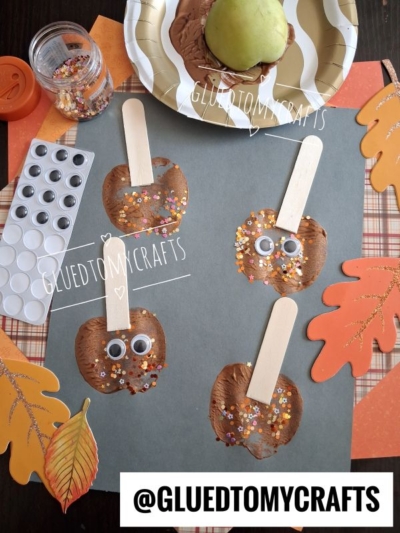 Four brown painted paper apples with googly eyes, craft sticks, and colorful glitter are arranged on dark paper with autumn leaves, a paint palette, and a plate with a real green apple in the background.