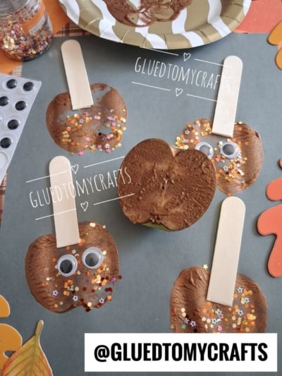 Four brown apple crafts with popsicle sticks, googly eyes, and glitter are arranged on a table with a real sliced apple, craft supplies, and orange decorations. The text @GLUEDTOMYCRAFTS is visible at the bottom.