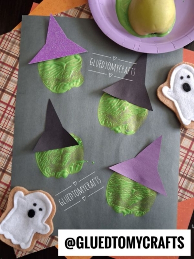 A craft sheet shows a charming apple stamped craft, featuring green apple prints decorated as witches with black hats. Felt ghost cookies and Halloween-themed papers are nearby. @GLUEDTOMYCRAFTS watermark is visible.