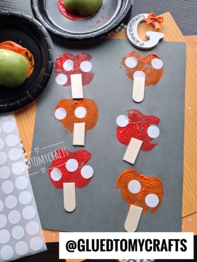 Gray paper shows six mushroom shapes made from orange and red paint with wooden sticks as stems and white paper circles as spots. This adorable APPLE STAMPED CRAFT scene also features craft supplies, including a green apple, plates, and a finished mushroom magnet.