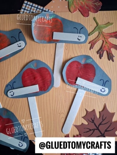 Paper crafts of smiling worms with red apples, created using an APPLE STAMPED CRAFT technique, painted on blue paper and attached to white popsicle sticks. The crafts are displayed on a wood-patterned surface with autumn leaf decorations. @GLUEDTOMYCRAFTS watermark appears.