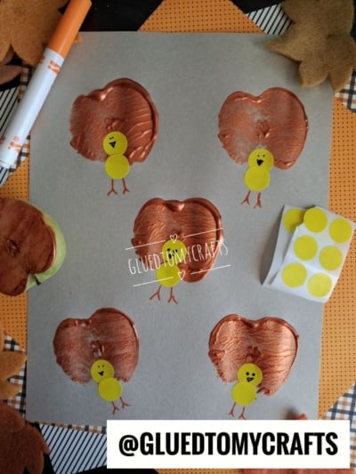 Five apple stamped turkeys made from copper-painted apple print bodies and yellow sticker heads, arranged on gray paper.