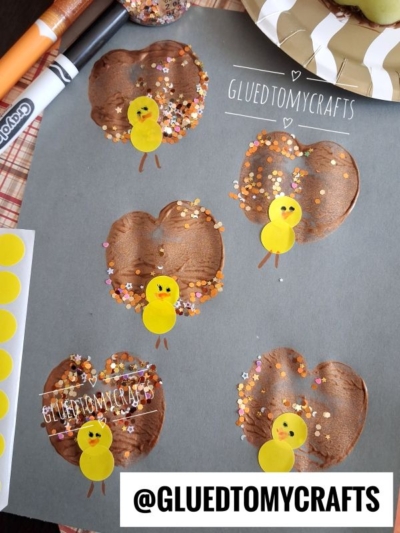 Brown apple prints on gray paper are decorated with yellow sticker chicks, glitter, and drawn legs. The words GLUEDTOMYCRAFTS and @GLUEDTOMYCRAFTS appear on the image. Markers and craft tools are nearby.