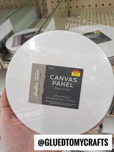 A hand holds a pack of round canvas panels, 6 inches in size, labeled Canvas Panel, priced at $2.49. Shelves with similar art supplies are visible in the background.