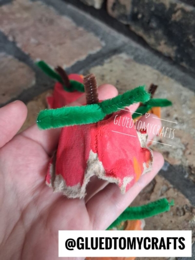 A hand holds a red-painted egg carton craft resembling an apple, with a brown pipe cleaner stem and green pipe cleaner as a leaf—making it a perfect idea for FALL CRAFTS FOR KIDS. The background shows a brick surface. Text: @GLUEDTOMYCRAFTS.