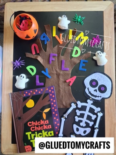 A Halloween-themed craft setup with a felt tree, colorful felt letters, ghost and skeleton cutouts, purple spiders, a pumpkin bucket, and the book Chicka Chicka Trick or Treat on a chalkboard—perfect inspiration for those looking for creative FALL THEMED SENSORY BIN IDEAS. @GLUEDTOMYCRAFTS.