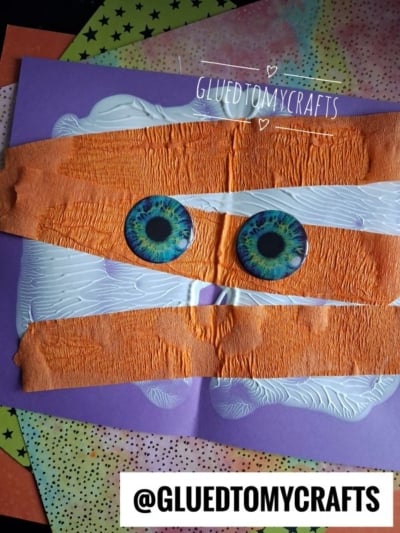 A kids paint splat mummy craft made with orange tape, two large green eyes, and white paint splatters on purple paper. Colorful star and dot papers are in the background. @GLUETOMYCRAFTS is written on the image.