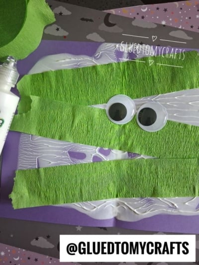 A spooky craft features green tissue strips, two large googly eyes, white glue, and a purple background with bats and stars. A label reads @GLUEDTOMYCRAFTS.