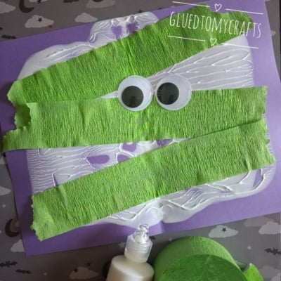 A kids craft project: green crepe paper strips and googly eyes glued on purple paper to resemble a mummy. Glue bottle and green crepe paper roll are nearby. @GLUEDTOMYCRAFTS is written at the bottom.