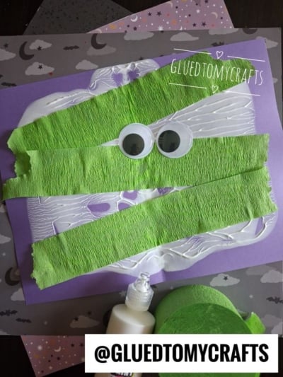 A kids craft project: green crepe paper strips and googly eyes glued on purple paper to resemble a mummy. Glue bottle and green crepe paper roll are nearby. @GLUEDTOMYCRAFTS is written at the bottom.