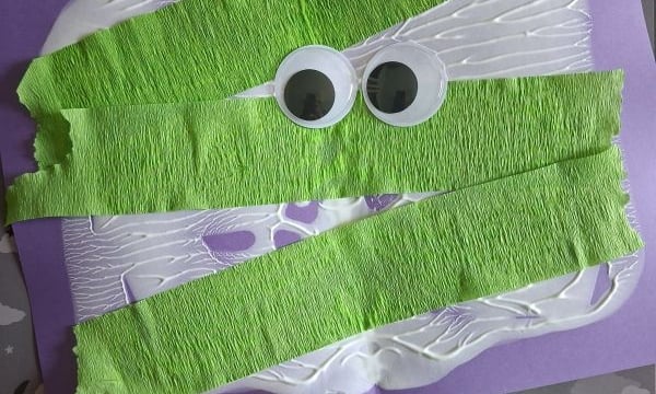 A kids craft project: green crepe paper strips and googly eyes glued on purple paper to resemble a mummy. Glue bottle and green crepe paper roll are nearby. @GLUEDTOMYCRAFTS is written at the bottom.