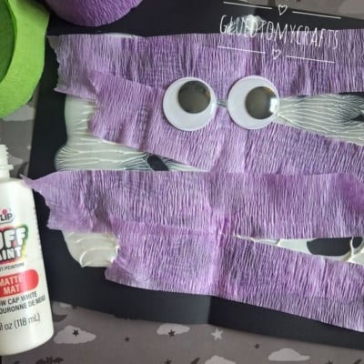 A Halloween craft with googly eyes and purple crepe paper strips glued to black paper, surrounded by craft supplies. The username @GLUEDTOMYCRAFTS is at the bottom.