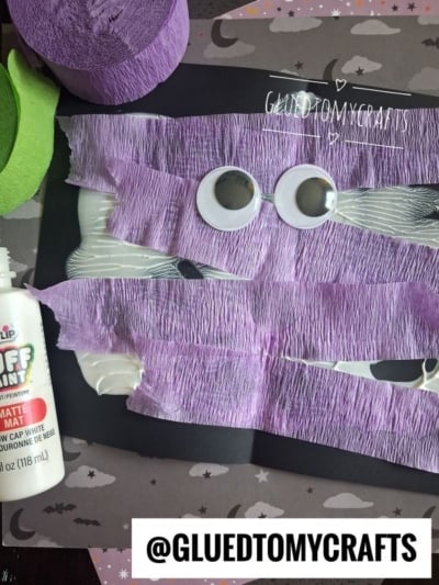 A Halloween craft with googly eyes and purple crepe paper strips glued to black paper, surrounded by craft supplies. The username @GLUEDTOMYCRAFTS is at the bottom.