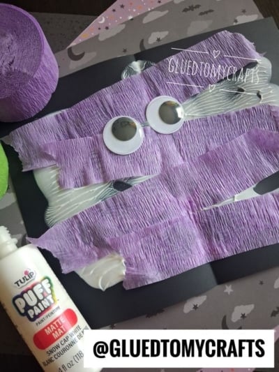 A mummy craft made with purple crepe paper, googly eyes, and a black background. A bottle of white puff paint and purple crepe paper roll are nearby. Halloween-themed paper with bats is underneath.