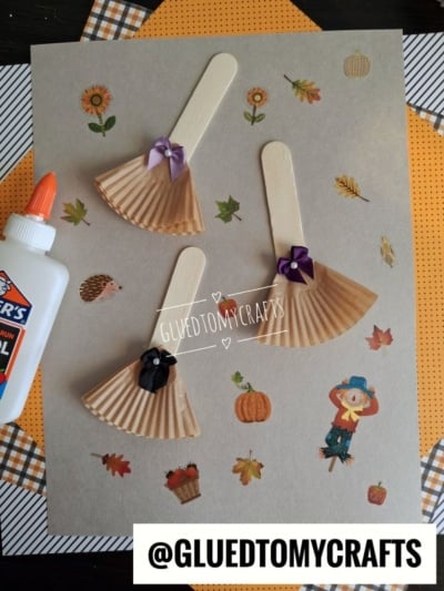 Three DIY cupcake liner broom crafts made from popsicle sticks and brown paper, decorated with small bows and placed on a background with fall-themed stickers.