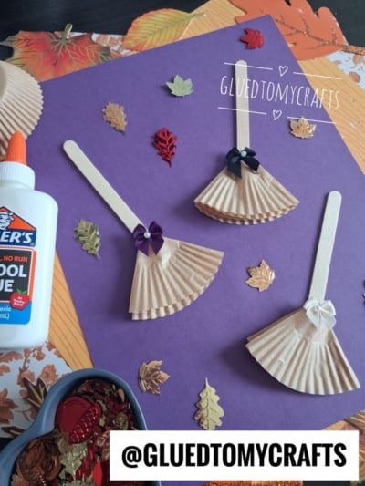 Three craft broomsticks made from popsicle sticks and folded paper, decorated with bows, are arranged on a purple background with autumn leaf cutouts. A bottle of glue and a tray of leaf-shaped confetti are nearby.