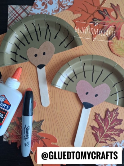 Two paper plate turkeys with brown heart-shaped faces, drawn eyes, and popsicle stick beaks are shown on a fall-themed background with leaves, glue, and a marker nearby—perfect for inspiring fun FALL CRAFTS FOR KIDS.