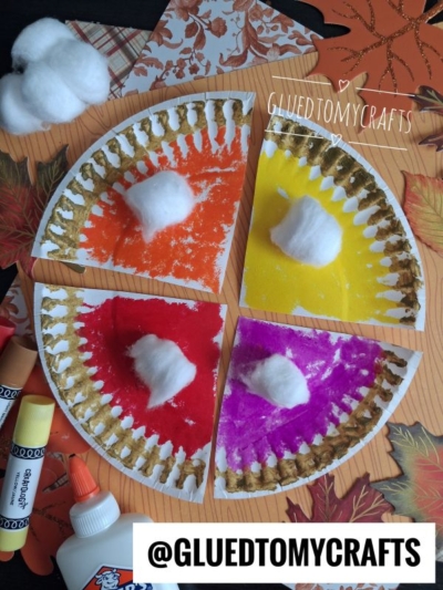 Four paper plate pie slices painted orange, yellow, purple, and pink with gold edges and a cotton ball on each, arranged on fall-themed paper. Craft supplies are nearby—making this a perfect idea for FALL CRAFTS FOR KIDS. Text: @GLUEDTOMYCRAFTS.