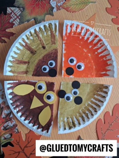 Four paper plate crafts painted as fall animals, including a lion, fox, owl, and mouse—each with googly eyes—sit on a table decorated with autumn leaves. These adorable FALL CRAFTS FOR KIDS showcase creativity and seasonal fun. The handle @GLUEDTOMYCRAFTS is visible at the bottom.