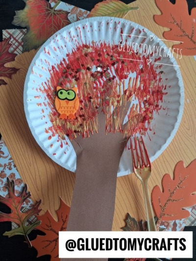 A delightful FALL CRAFTS FOR KIDS project, this paper plate craft features a fall tree with a brown paper hand as the trunk, vibrant red and orange leaves, and a charming little owl. A plastic fork with paint is placed beside it for added creativity. @GLUEDTOMYCRAFTS is written on the image.