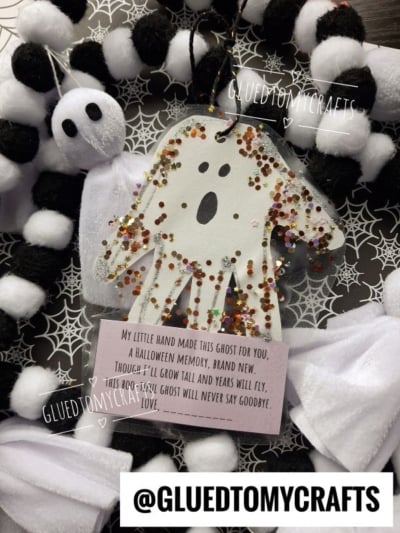 A handmade handprint ghost ornament with glitter and a poem hangs on a black-and-white pompom wreath. A small felt ghost and spiderweb-patterned background are also visible. Text reads @GLUEDTOMYCRAFTS.
