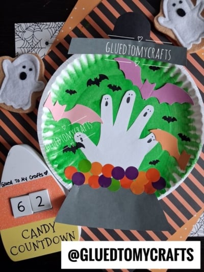 A Halloween craft scene with paper bats, a handprint ghost, and colorful dots in a cauldron on a green background, plus a candy countdown and ghost decorations—perfect inspiration for Halloween Crafts for Kids. @GLUEDTOMYCRAFTS is labeled.