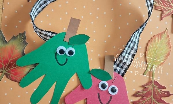 Two paper handprint cutouts, one green and one red, with googly eyes and smiles, hang from a ribbon on a fall-themed background with leaves. The ribbon reads, Love you bunches & bushels..