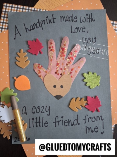 A handmade craft with a floral handprint cutout shaped like a hedgehog, surrounded by paper leaves, handwritten text, a gold marker, and autumn-patterned papers in the background—perfect inspiration for FALL CRAFTS FOR KIDS.