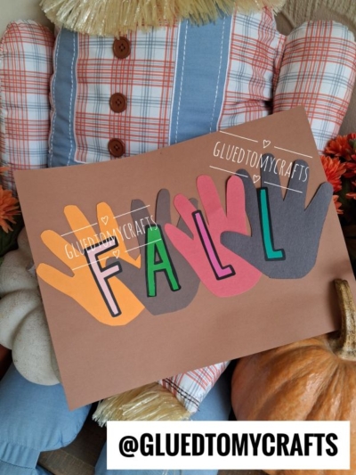 A fall craft features four colorful paper handprints spelling FALL, held by a scarecrow in plaid beside pumpkins and autumn leaves—an ideal project for FALL CRAFTS FOR KIDS. @GLUEDTOMYCRAFTS is written at the bottom.