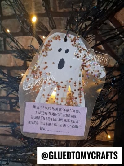 A paper ghost craft with glitter and a poem hangs on a black tree. The poem is about a handprint ghost that will never say goodbye. The Instagram handle @GLUEDTOMYCRAFTS is at the bottom.
