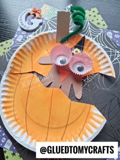 A paper plate pumpkin craft with an orange cutout, googly eyes, cupcake liner glasses, pipe cleaner stem, and paper hand nose on a spiderweb background. @GLUEDTOMYCRAFTS is written at the bottom.