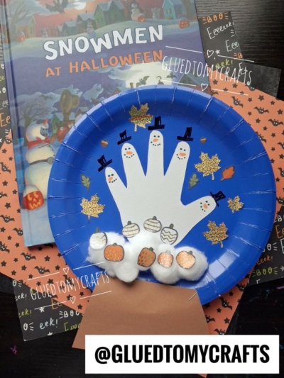 Blue plate with a white handprint snowman snow globe craft, decorated with hats and pumpkins, cotton ball snow, and gold leaves, on Halloween-themed paper with the Snowmen at Halloween book in the background.