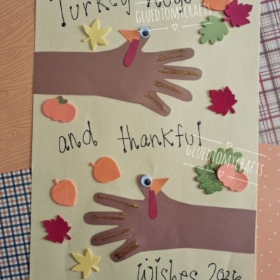 Handprint turkey crafts with paper hands decorated as turkeys, surrounded by leaf and pumpkin cutouts. Text reads Turkey hugs and thankful wishes 2026. @GLUEDTOMYCRAFTS is at the bottom.