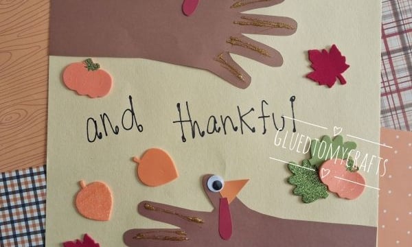 Handprint turkey crafts with paper hands decorated as turkeys, surrounded by leaf and pumpkin cutouts. Text reads Turkey hugs and thankful wishes 2026. @GLUEDTOMYCRAFTS is at the bottom.